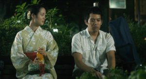 Trecho do filme Summer Blooms, em cena noturna, mostra uma mulher em trajes típicos conversando com um homem