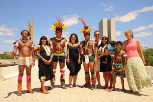 Dez pessoas posam para foto, sendo 5 adultos e duas crianças indígenas. Ao fundo, edifícios da Esplanada de Brasília