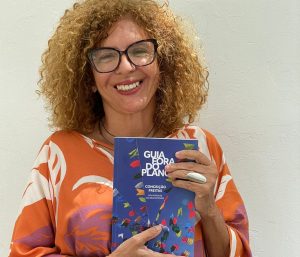 Conceição Freitas vestida de blusa alaranjada, sorrindo, com óculos, segurando o livro Guia Fora do Plano