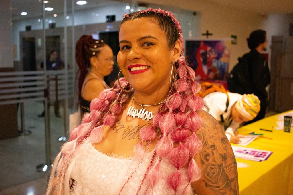 Mulher de cabelo rosa e tranças, sorri para a câmera