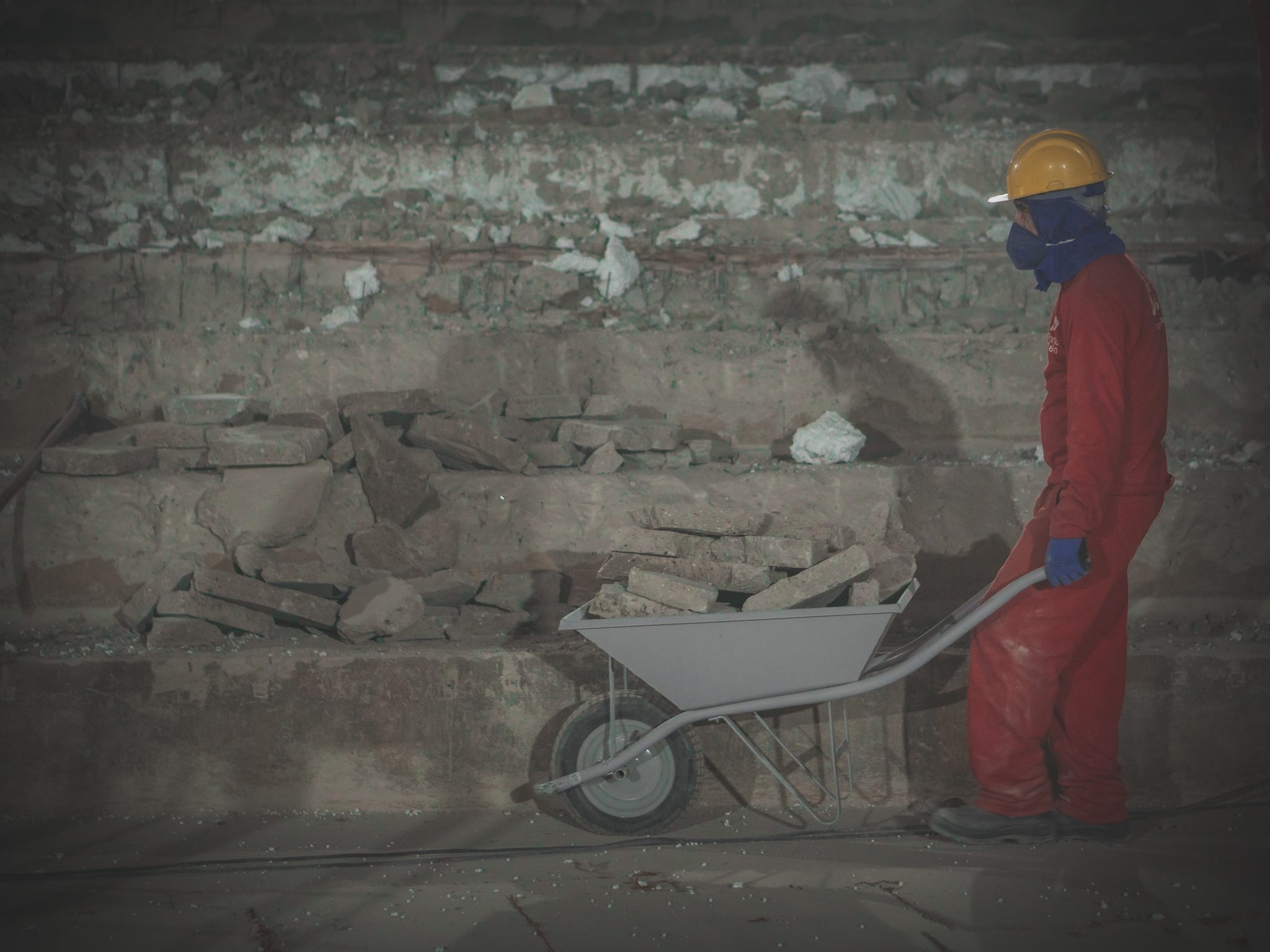 Home de vermelho e capacete de obras amarelo caminha num canteiro de obras empurrando um carrinho de mão