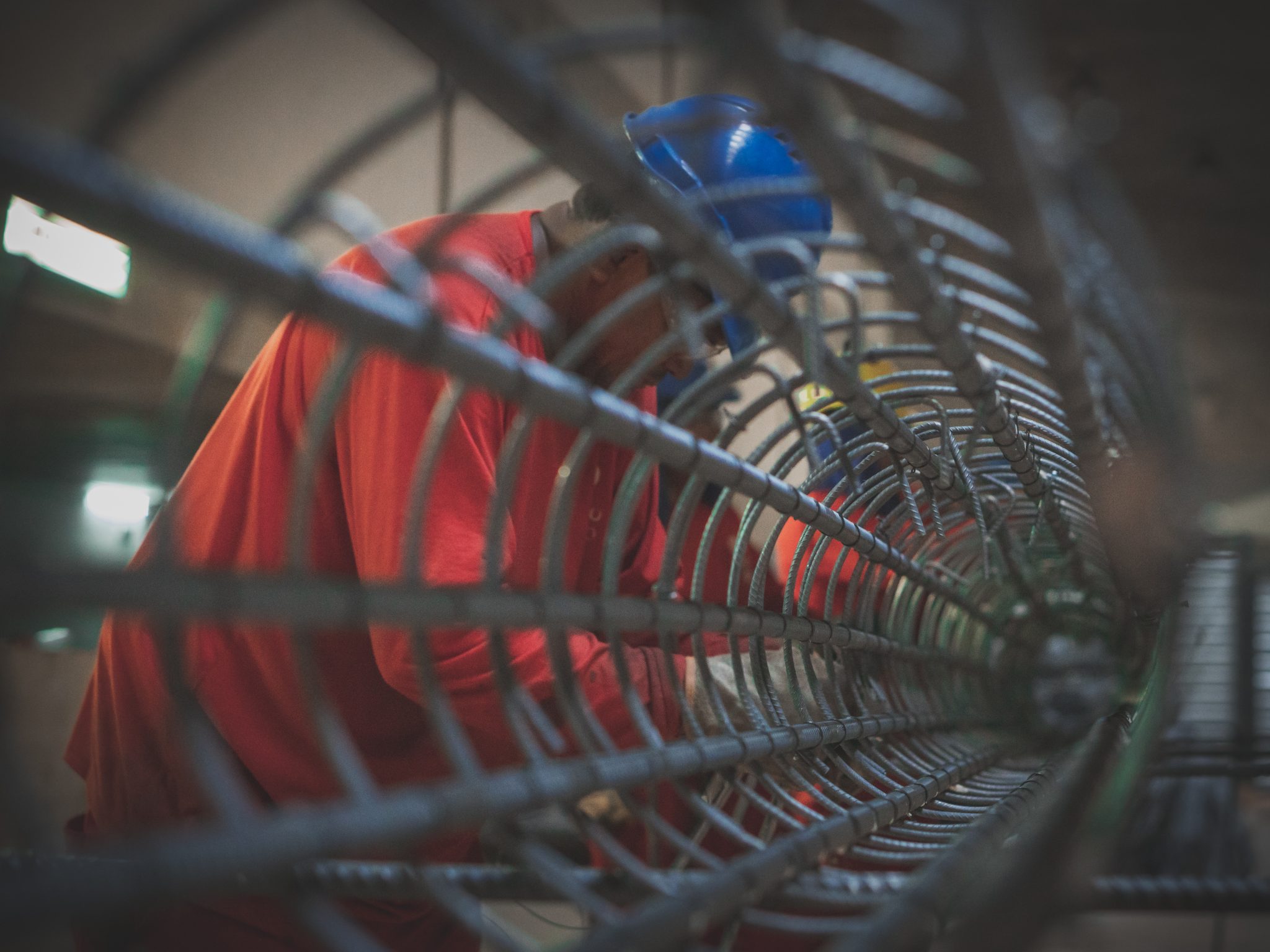 Homem vestido de vermelho e usando capacete azul de obras trabalhando numa estrutura de ferro