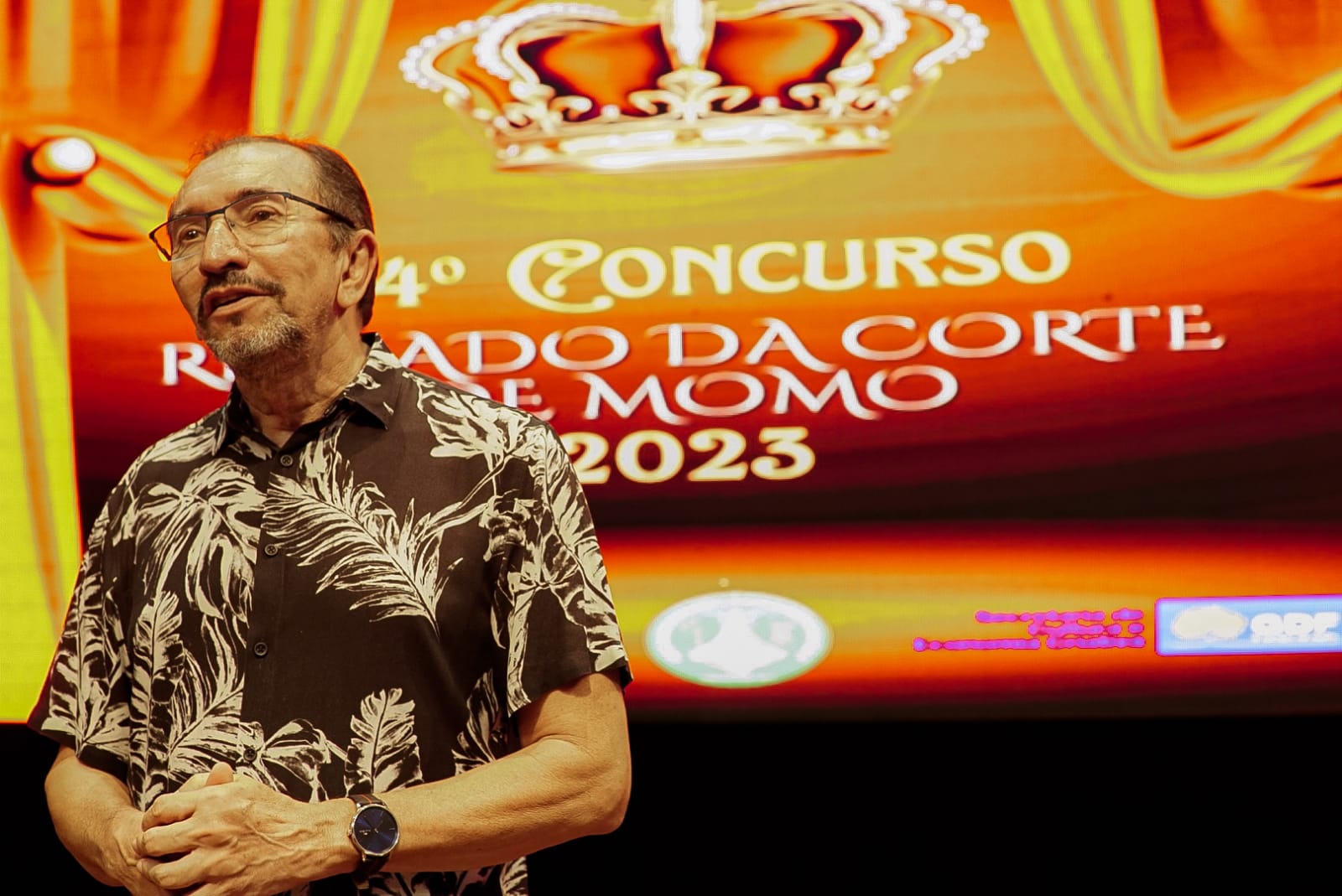 Homem vestindo camisa com estampa colorida, as duas mãos apoiadas uma na outra com um painel ao fundo