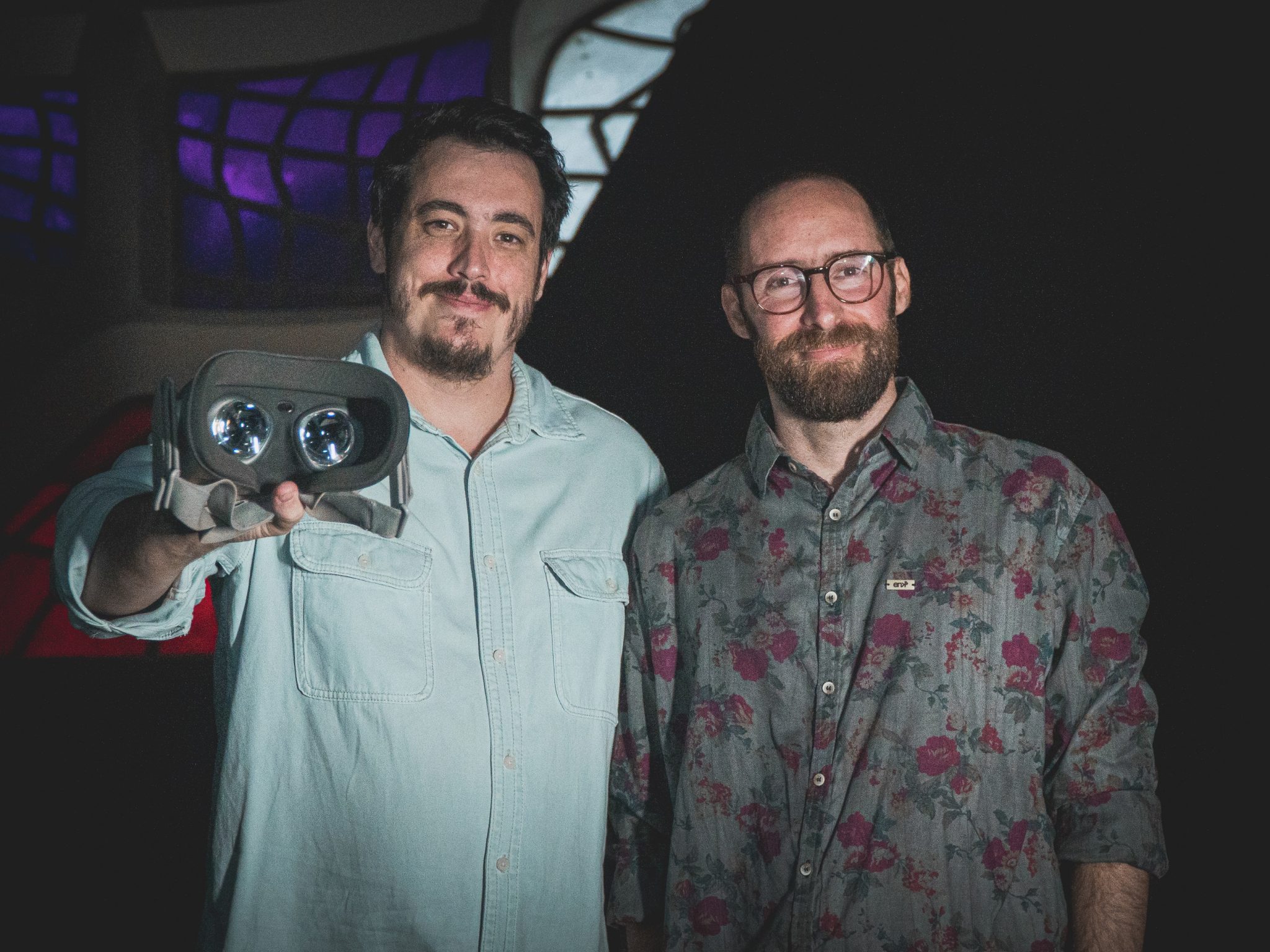 Dois homens olhando para a câmera, um deles segura um óculos de realidade virtual
