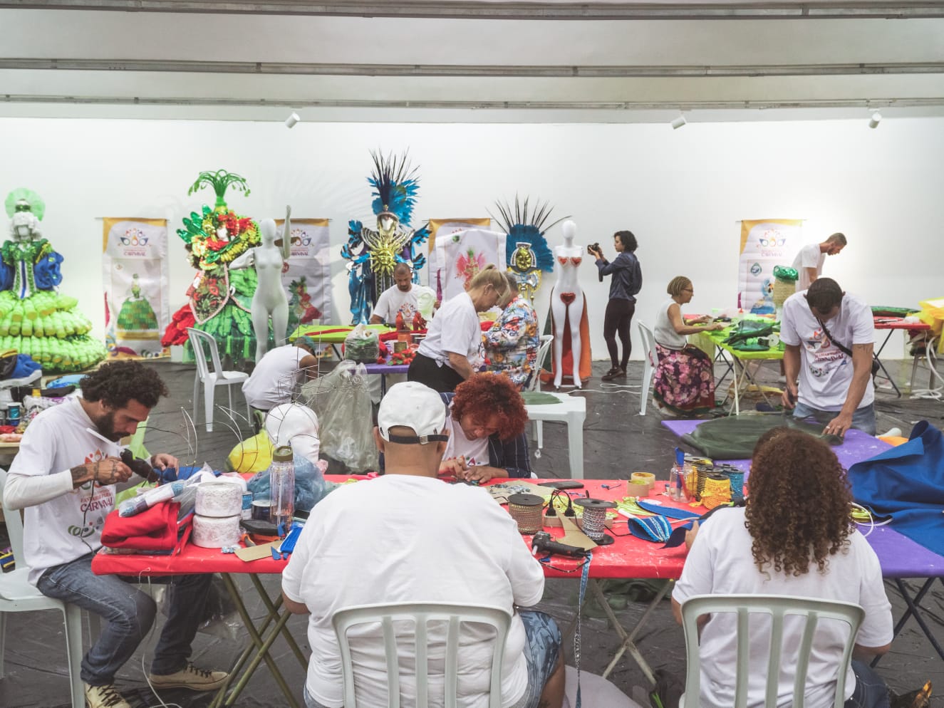 Galpão da Escola de Carnaval, com diversas pessoas produzindo entre adereços, tecidos e fantasias