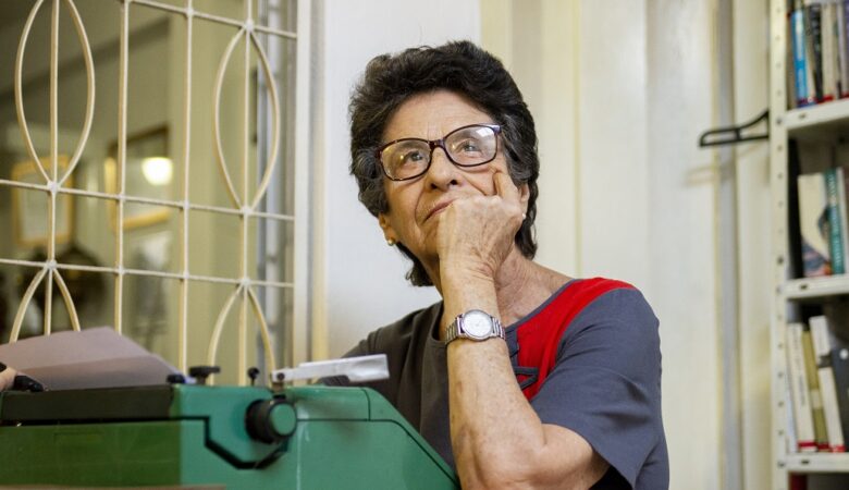 Mulher de óculos, em frente a uma máquina de escrever, coloca a mão no queixo
