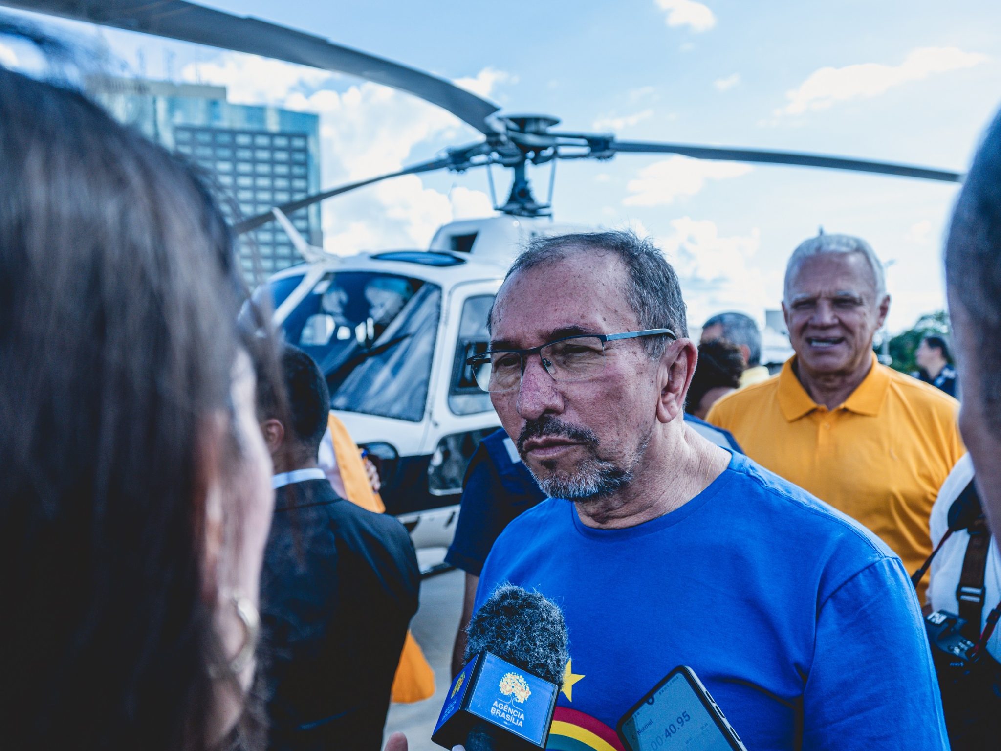Secretário Bartolomeu, de camisa azul e óculos, dá entrevista. Ao fundo, aparece um helicóptero