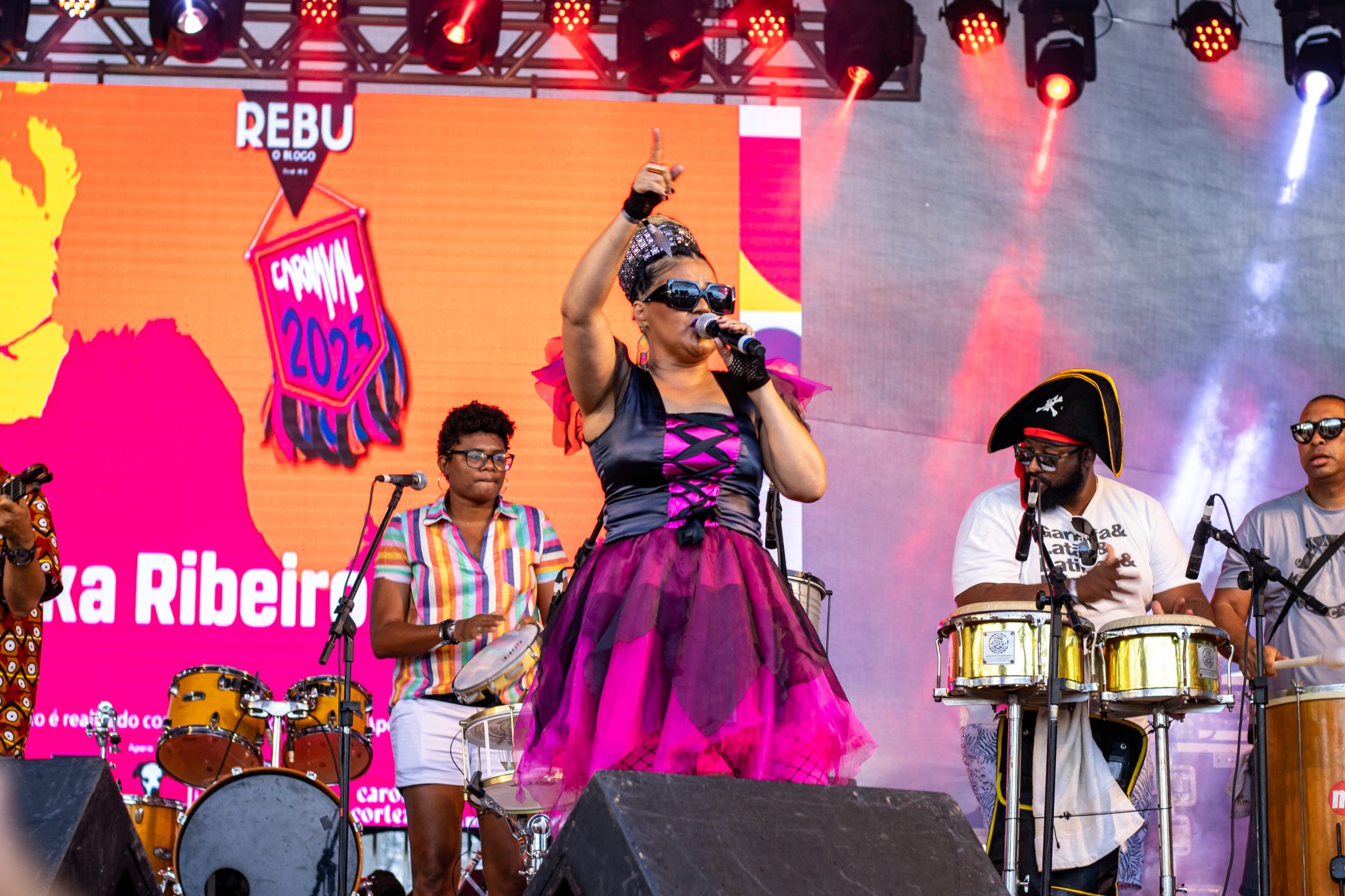 Imagem da vocalista no palco com vestido rosa e músicos ao fundo