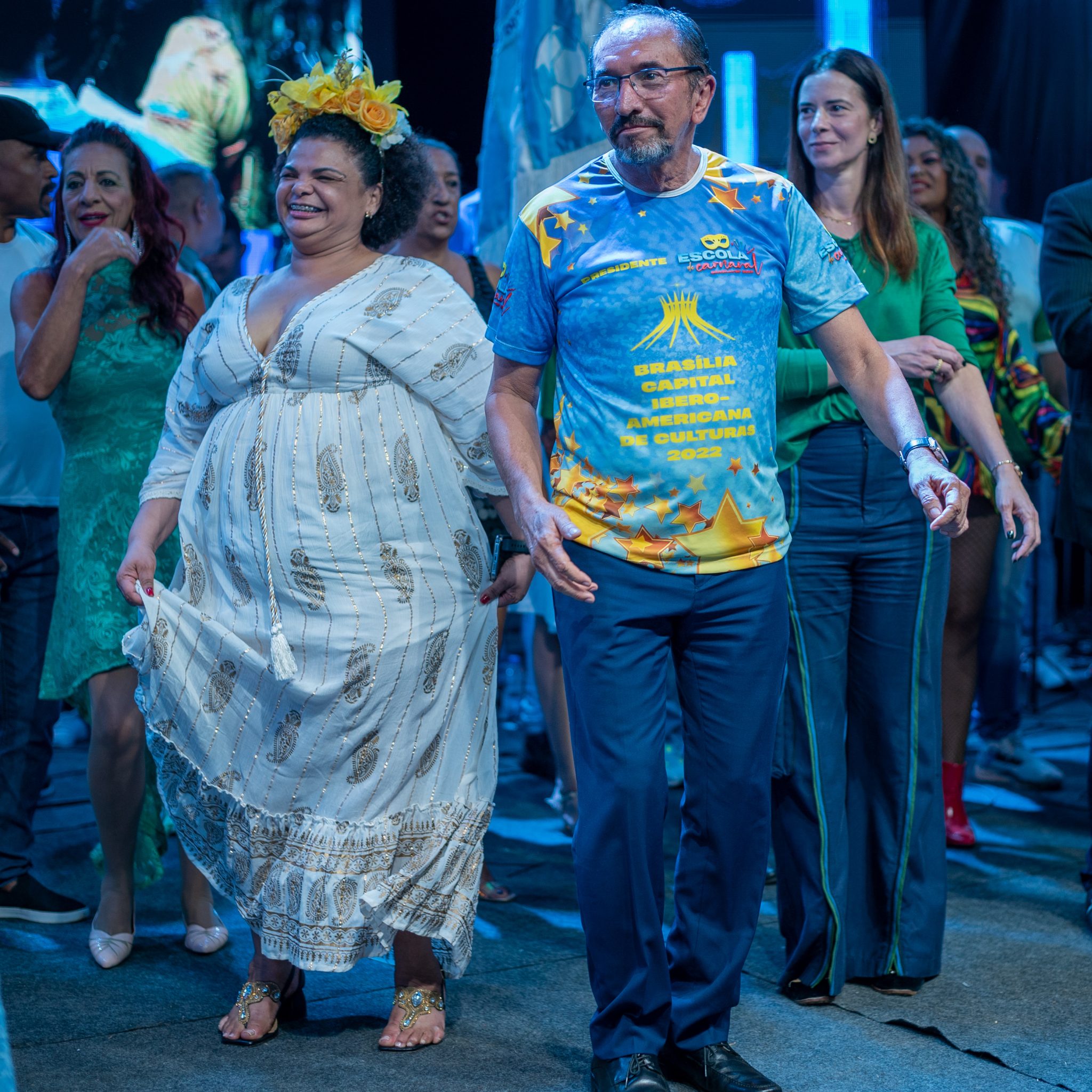 Subsecretária de Difusão e Diversidade, de vestido branco e arranjo de flores na cabeça, acompanhada do secretário de cultura, de camiseta da Escola de Carnaval