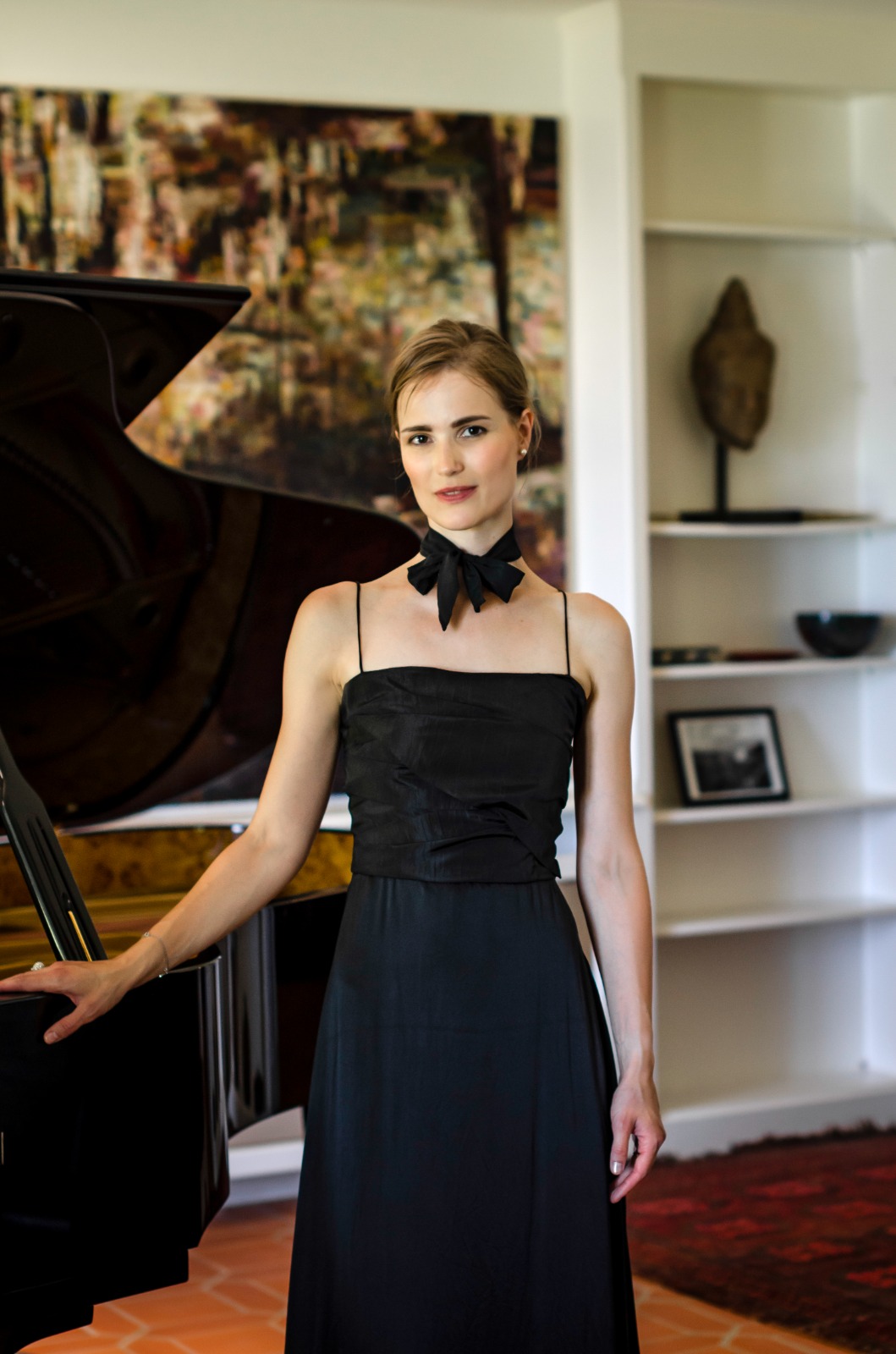 Mulher branca, de vestido preto e laço no pescoço, em frente a um piano