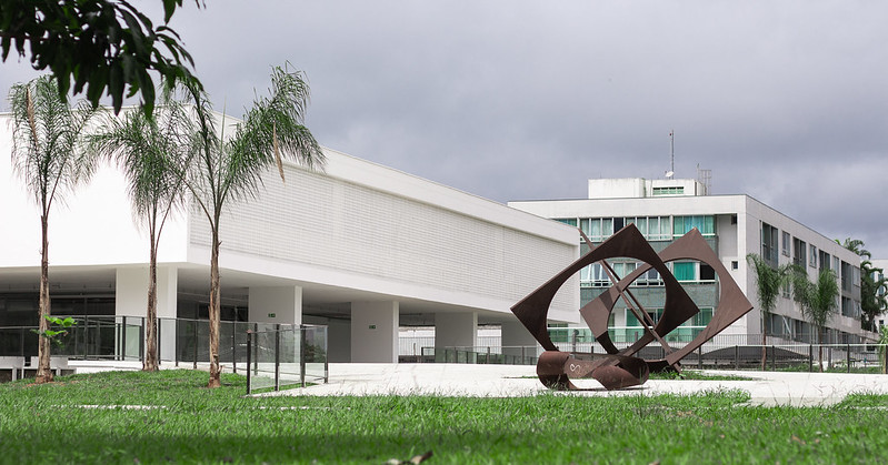 Fachada externa do MAB, edifício modernista branco sob pilotis. Em primeiro plano aparece um jardim e uma obra de arte