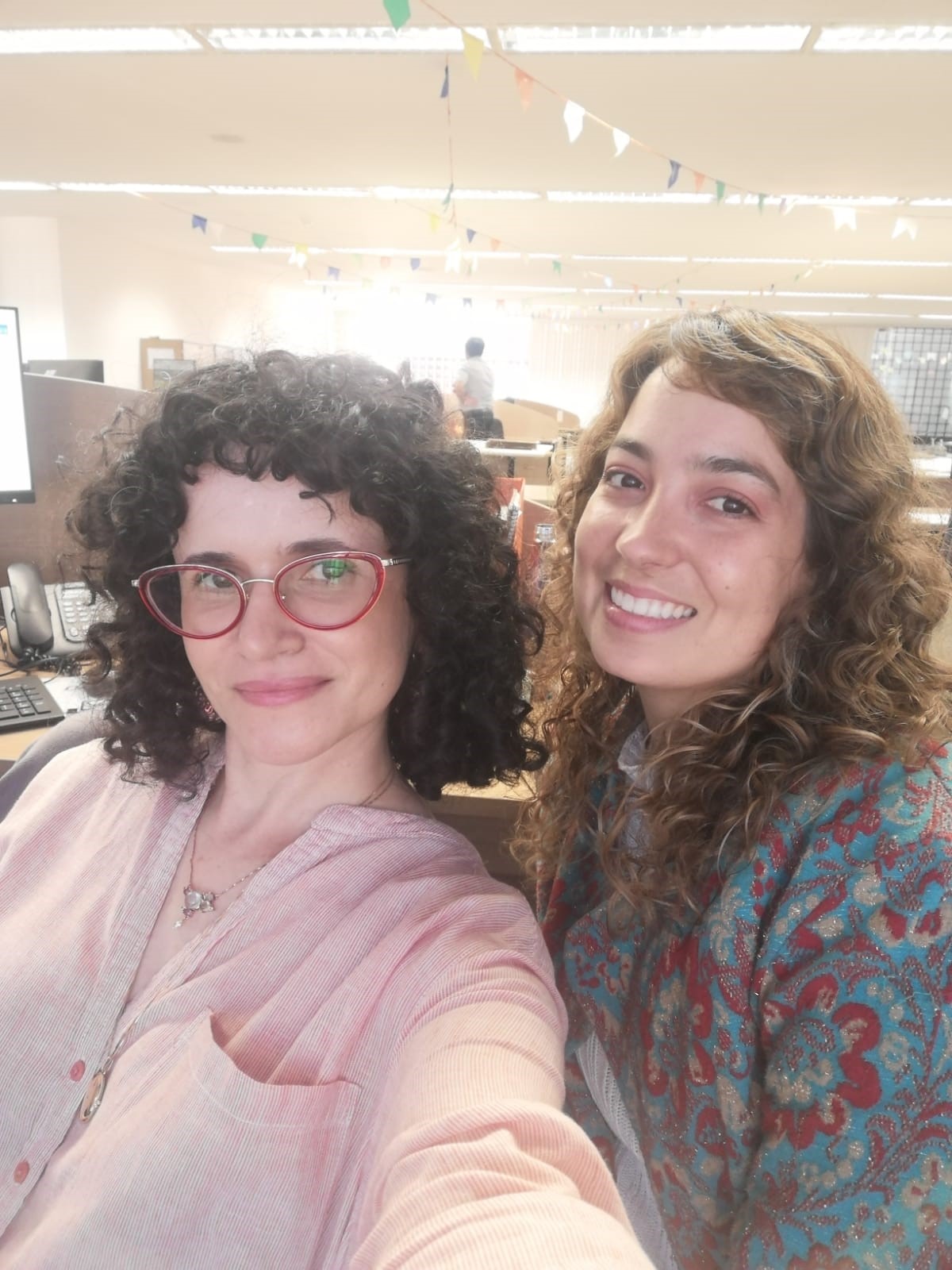 Foto das servidores Fátima, de cabelo preto e óculos, ao lado da colega Mariah. de cabelos longos castanhos e blusa estampada 