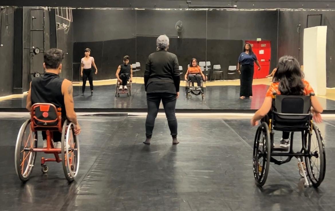 Foto de sala no Centro de Dança, com parede de espelho, dois cadeirantes nas pontas e uma idosa ao centro