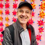 Foto de Sérgio Bacelar, Criador do Movimento Internacional de Dança, usando blusa golo polo cinza, com casaco e boné pretos, usando óculos, sorrindo, com um fundo rosa