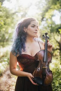 Violinista, mulher branca de cabelos coloridos segura o violino. Ao fundo, uma paisagem natural