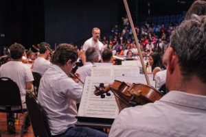 Músicos da Orquestra Sinfônica, todos de camiseta branca, se apresentam no Teatro Plínio Marcos