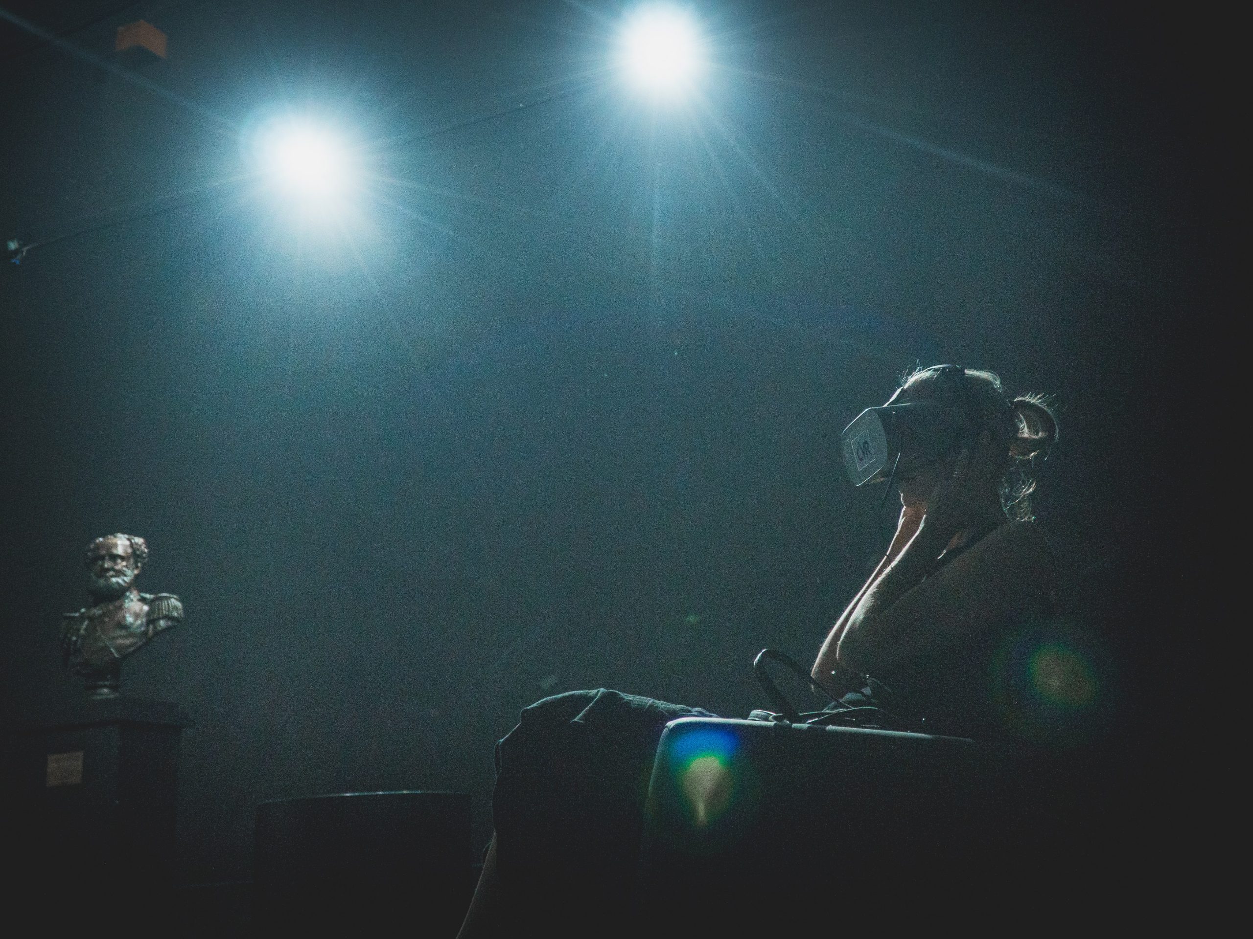 Em sala escura, com duas luzes superiores, mulher sentada usa óculos de realidade virtual